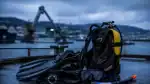 Imagen de equipo de buceo listo para inmersión en un puerto con fondo de maquinaria y agua. - Conjunto de equipo de buceo completo, incluyendo máscara, tanque y escarpines, preparado para una inmersión en puerto con vista al mar y maquinaria industrial.