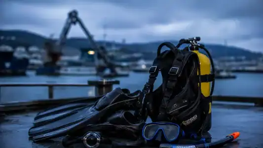 Conjunto de equipo de buceo completo, incluyendo máscara, tanque y escarpines, preparado para una inmersión en puerto con vista al mar y maquinaria industrial.